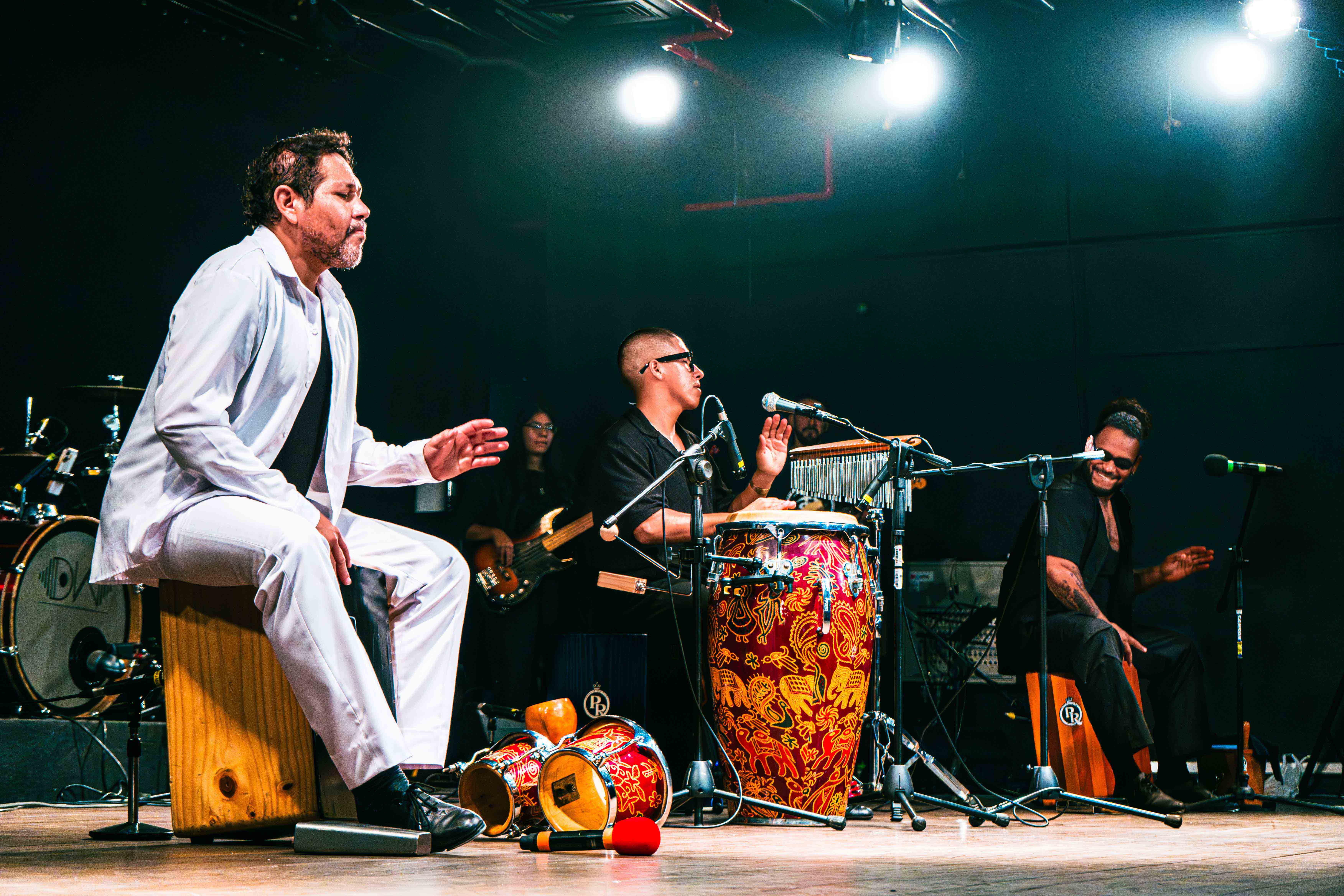 Foto que muestra en un escenario a tres músicos percusionistas: dos de ellos tocan cajón y el del medio toca una tumbadora. Los tres tienen ...